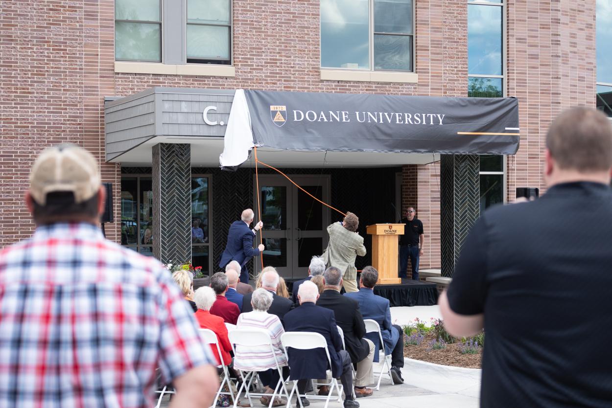 Dr. Roger Hughes (left), president of Doane University, and Roger Sheffield, former vice president of institutional advancement at Doane and founder and CEO of Aspen Philanthropy Group, take hold of two orange ropes to pull a banner and officially reveal C. Donald Brodie Hall.