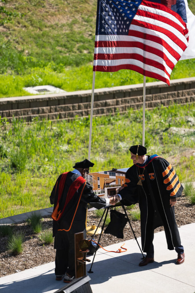 Rev. Dr. Robert L. Polk, a 1952 graduate of Doane University, shakes hands with Doane President Dr. Roger Hughes after they revealed a rendering of the Robert L. Polk Open Air Theatre. The naming of the theatre was celebrated during Doane’s baccalaureate ceremony, held in the theatre between the university’s C. Donald Brodie Hall and the Chab Weyers Education & Hixson Lied Art Building.